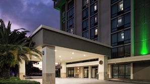 A hotel entrance with a covered walkway, palm trees, and a large building with multiple windows. - Holiday Inn Miami - International Airport by IHG (Miami Springs)