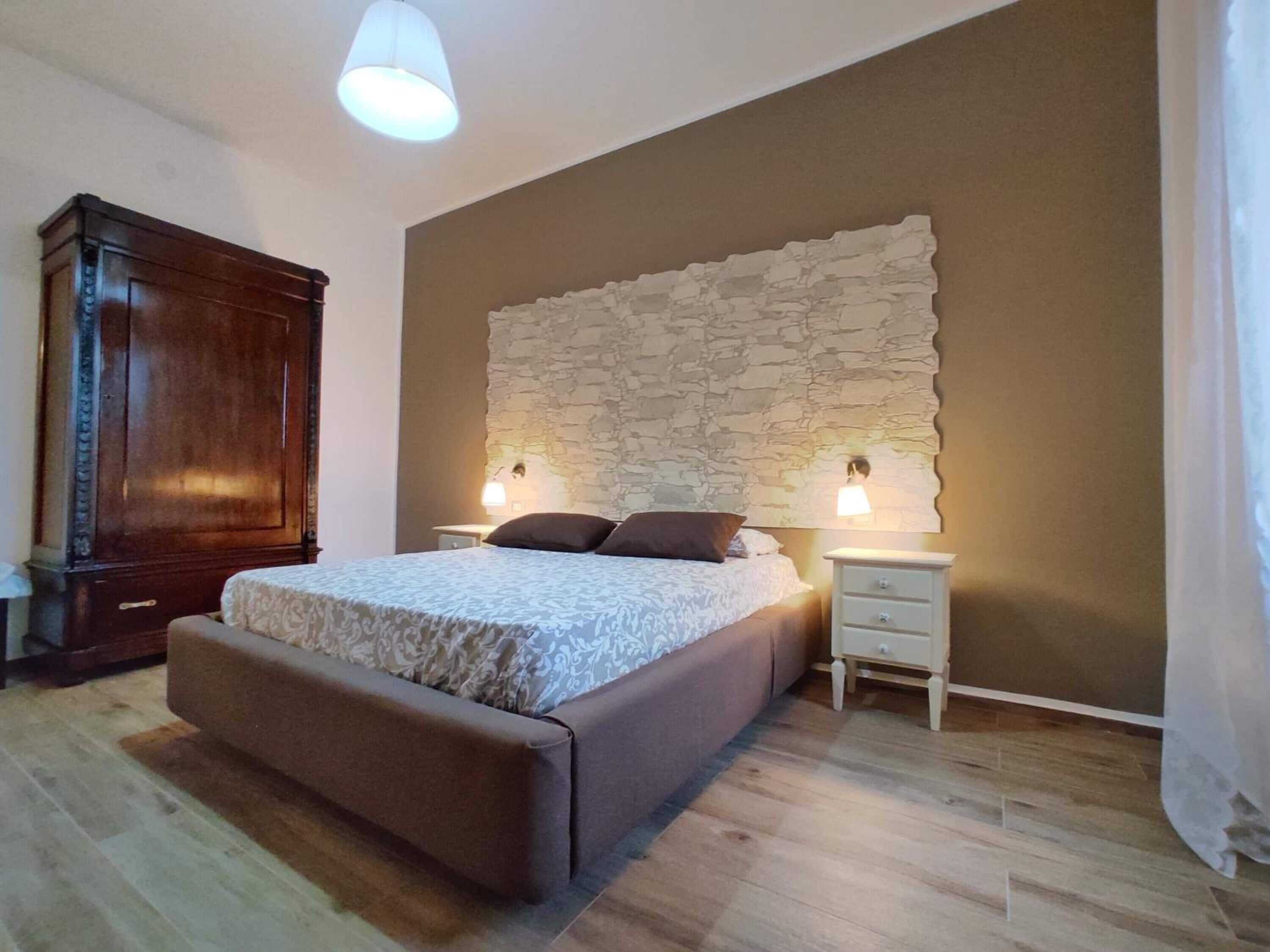 A bedroom with a large bed, a wooden wardrobe, a stone accent wall, and two bedside lamps.