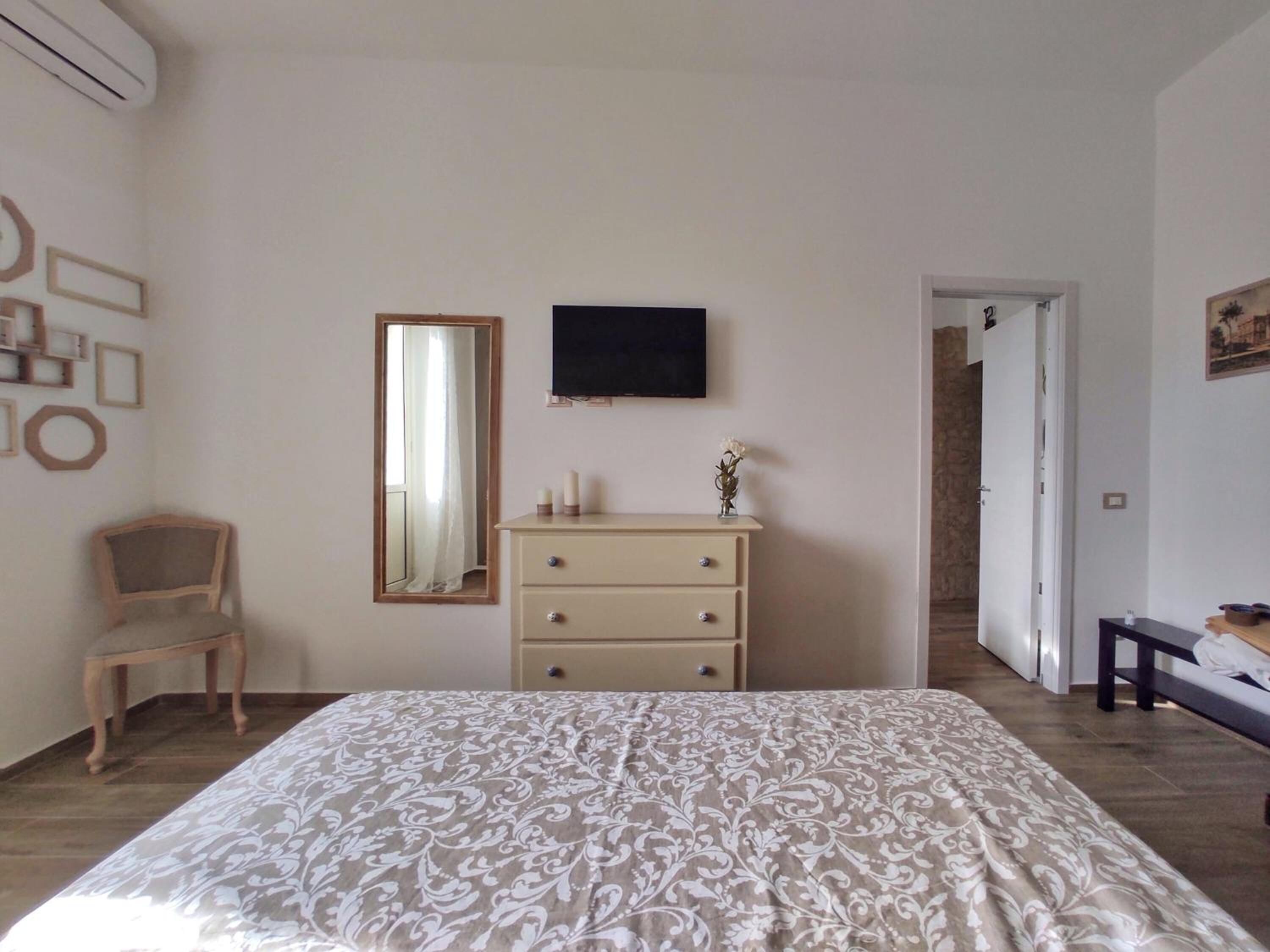 Un dormitorio con cama, cómoda, silla, espejo y televisor en la pared.