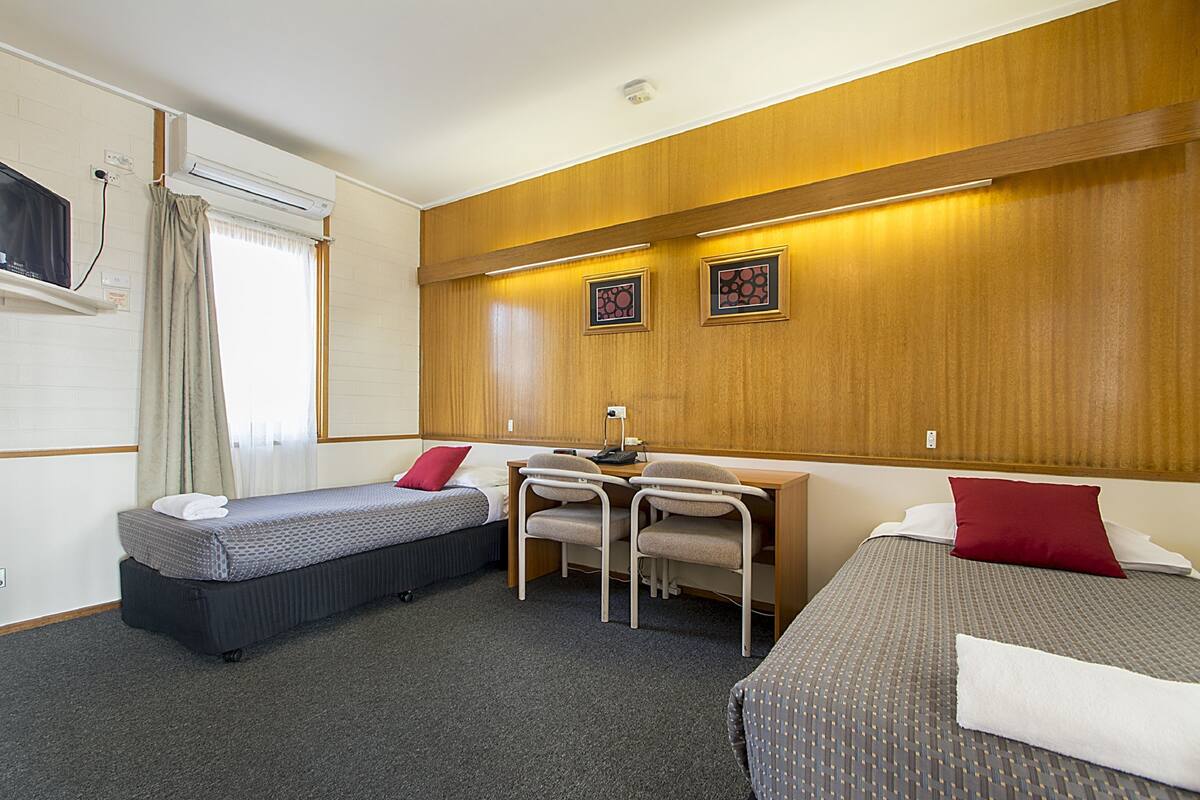 Habitación de hotel con dos camas individuales, un escritorio con dos sillas, un televisor y una ventana con cortinas.