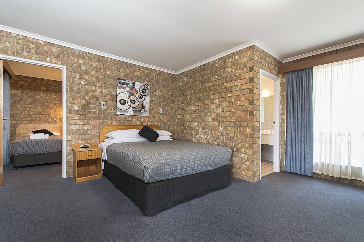 Un dormitorio con pared de ladrillo, cama con colcha gris y negra, mesita de noche y puerta que da a otra habitación.