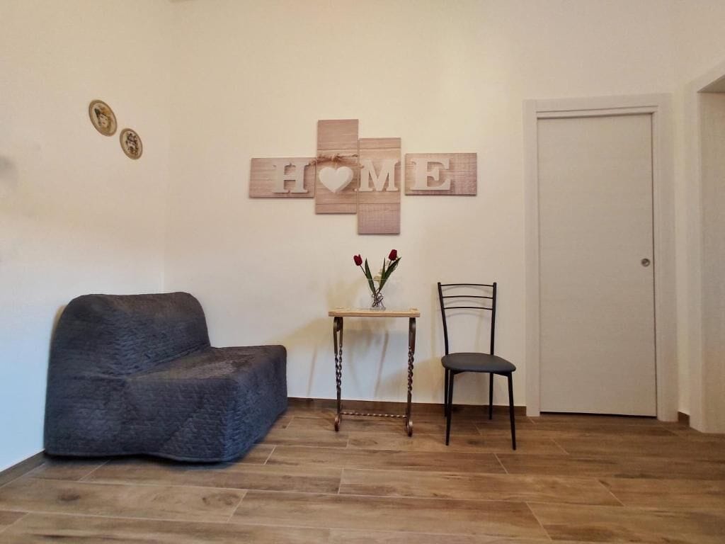 Une pièce avec un canapé gris foncé, une petite table en bois sur laquelle est posé un vase de fleurs, une chaise noire et un panneau mural portant l’inscription « HOME » avec un symbole en forme de cœur.