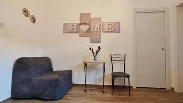Une pièce avec un canapé gris foncé, une petite table en bois sur laquelle est posé un vase de fleurs, une chaise noire et un panneau mural portant l’inscription « HOME » avec un symbole en forme de cœur.