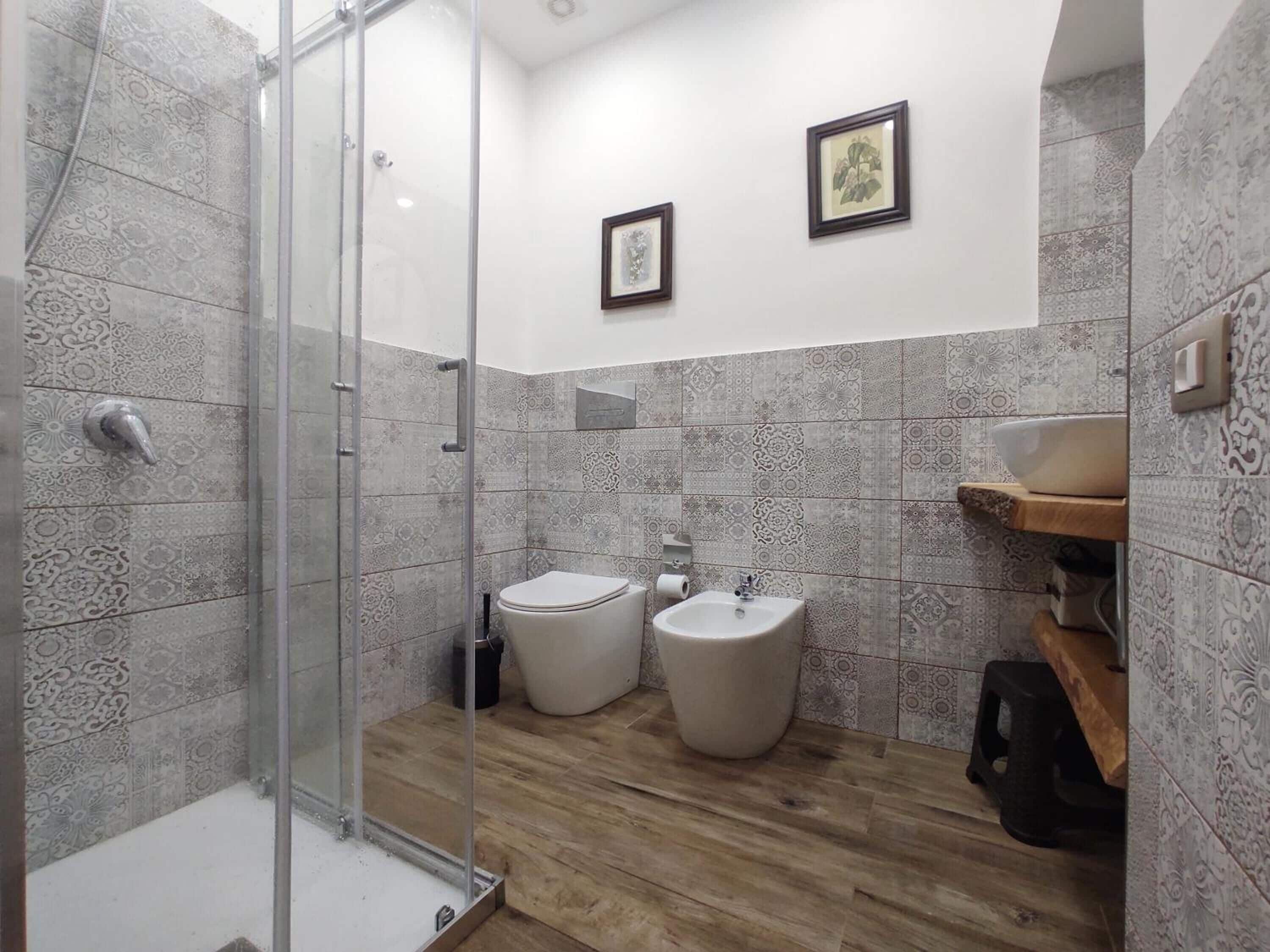 Een badkamer met patroontegels, een glazen douchecabine, een toilet, een bidet en een wastafel met een houten plankje.