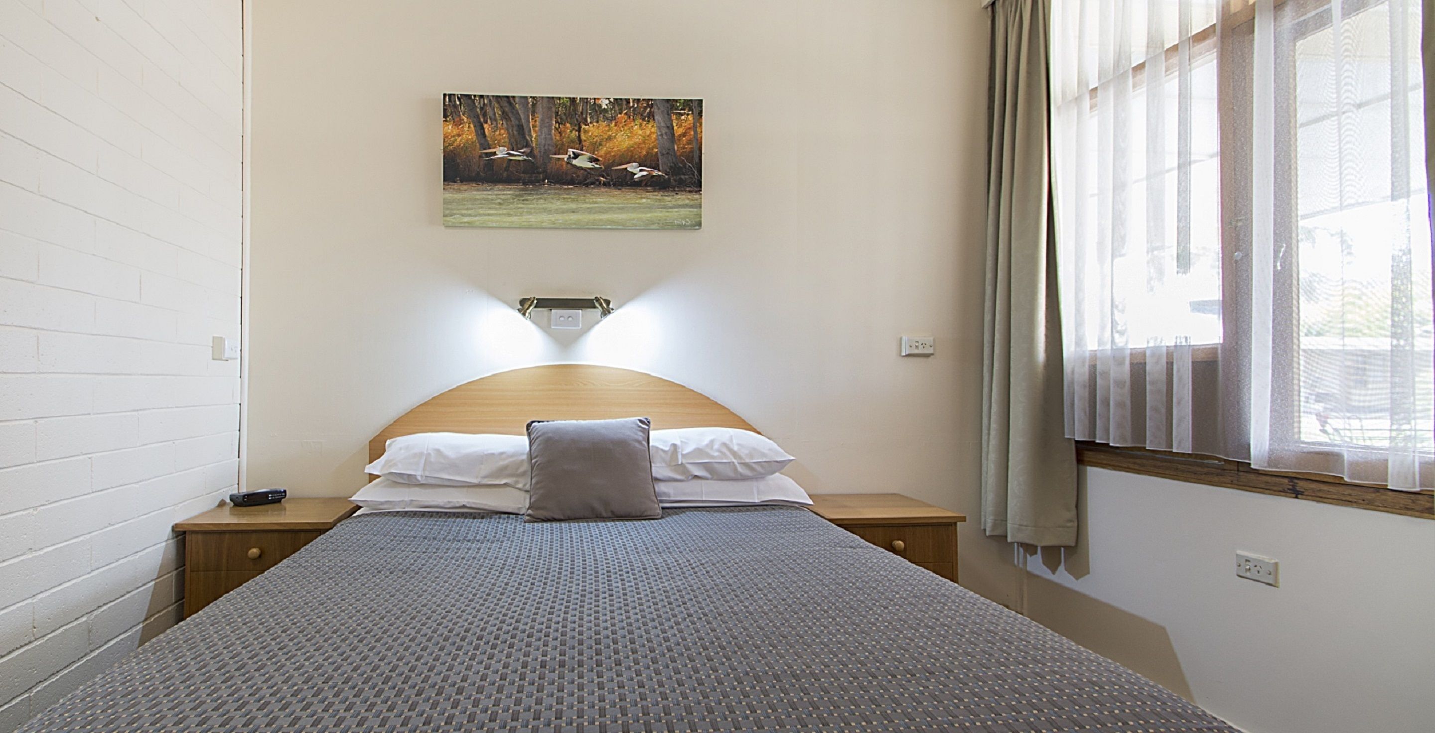 A neatly made bed with a patterned bedspread, a pillow, and a bedside table with a remote control. A painting of birds in flight hangs on the wall above the bed.