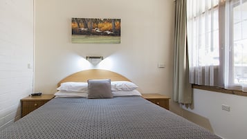 Un lit bien fait, recouvert d’une couvre-lit à motifs, avec un oreiller et une table de chevet sur laquelle se trouve une télécommande. Un tableau représentant des oiseaux en vol est accroché au mur, au-dessus du lit.