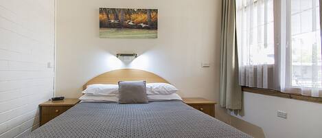A neatly made bed with a patterned bedspread, a pillow, and a bedside table with a remote control. A painting of birds in flight hangs on the wall above the bed.