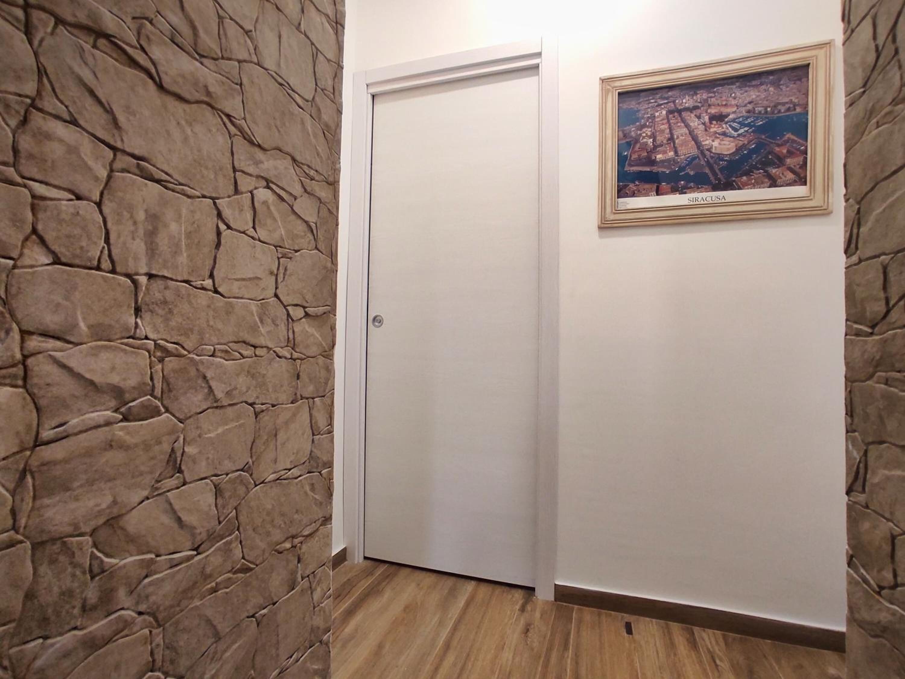 A hallway with stone walls, a wooden floor, a white door, and a framed picture of Siracusa.