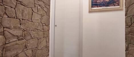 A hallway with stone walls, a wooden floor, a white door, and a framed picture of Siracusa.