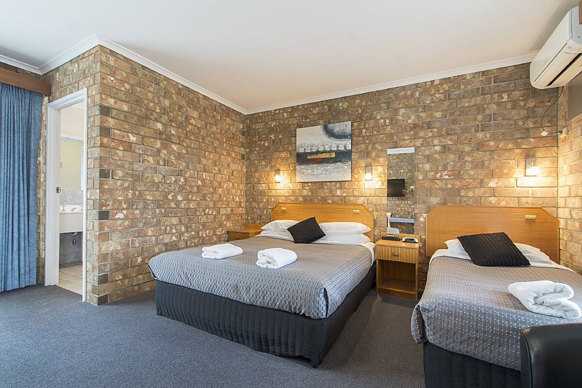 Habitación de hotel con dos camas, una pared de ladrillo, un cuadro y un baño visible a través de una puerta abierta.