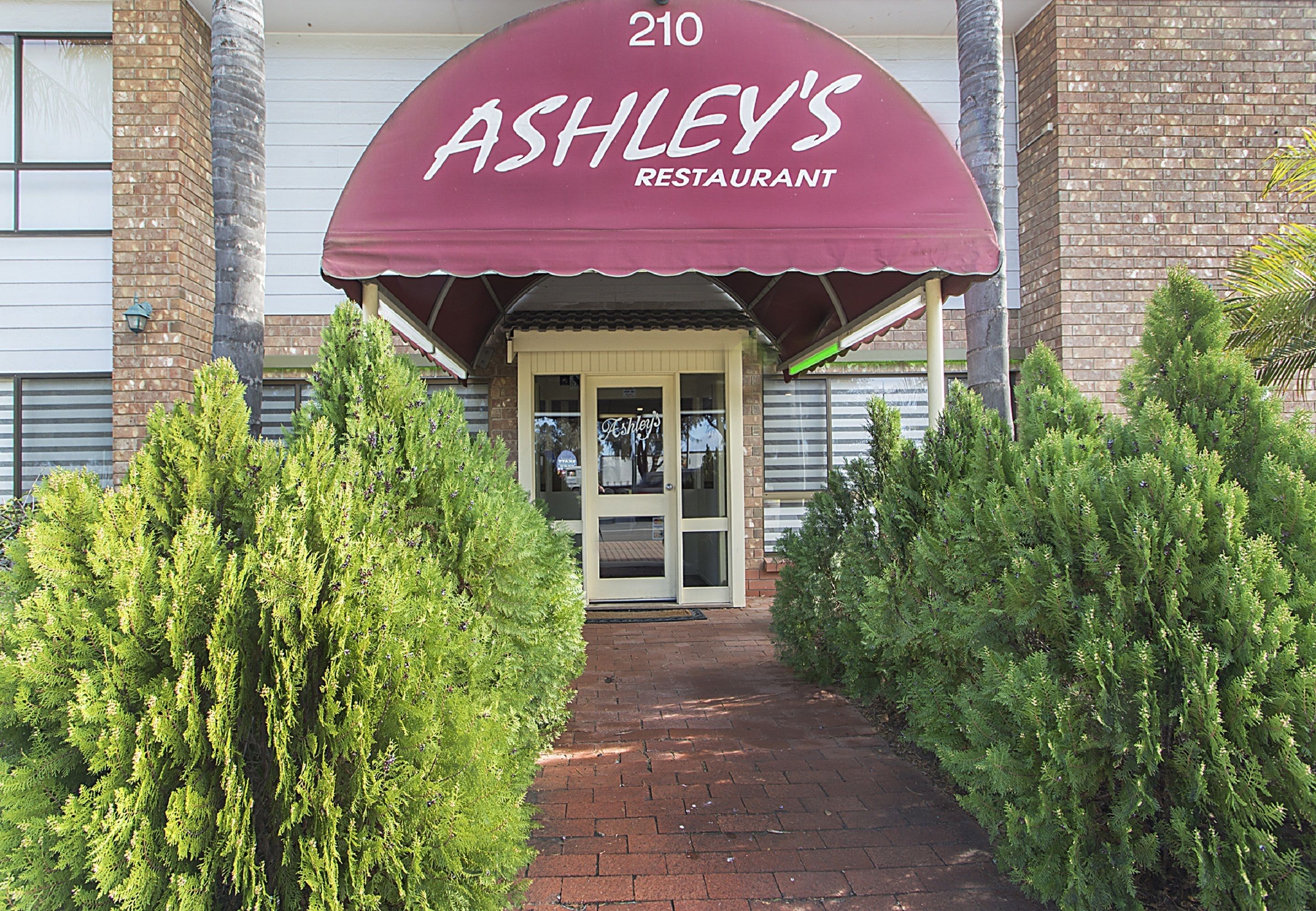 The entrance to Ashley's Restaurant, featuring a brick facade, a red awning with the restaurant's name, and a pathway flanked by green shrubs.