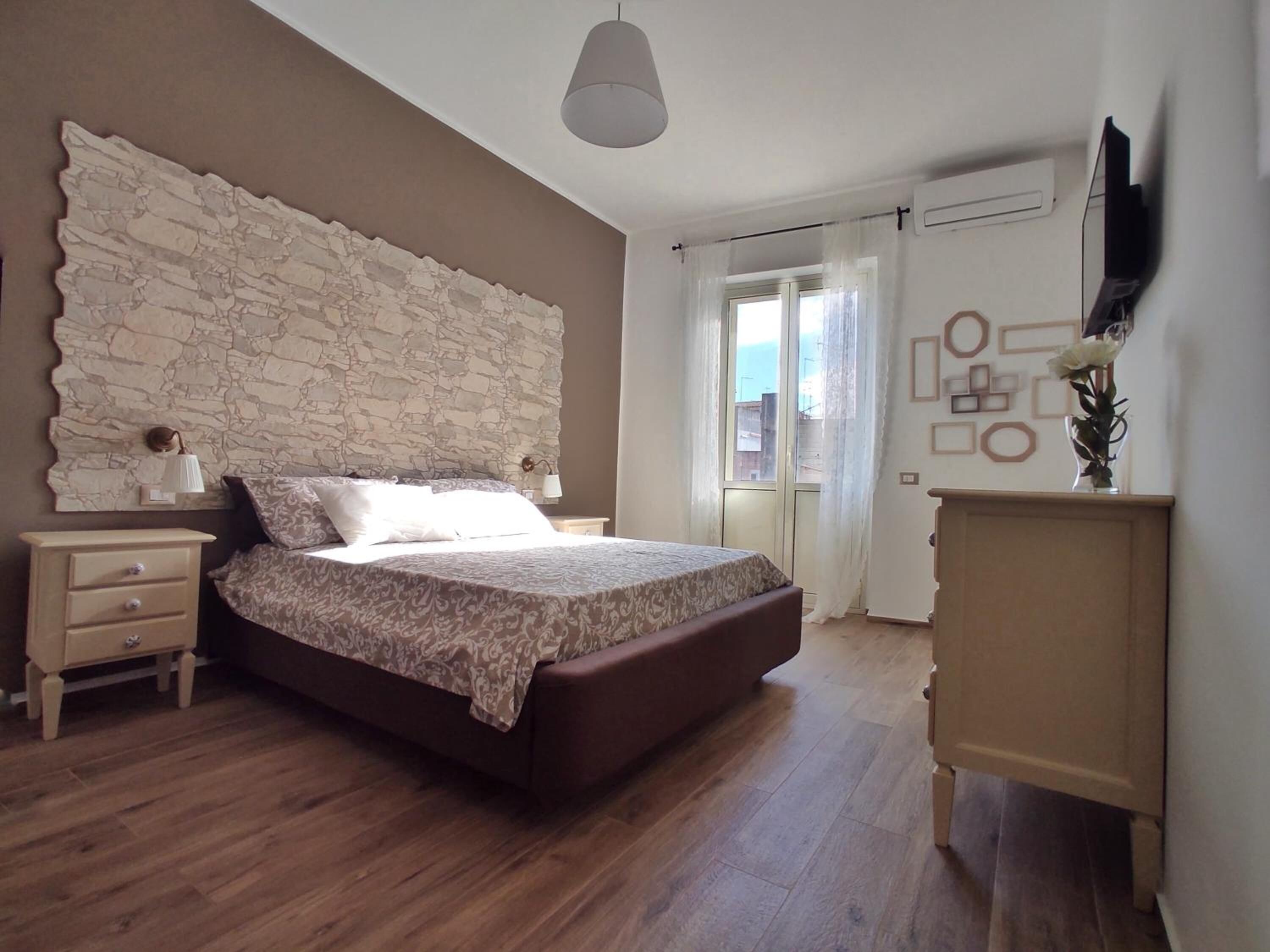 Een slaapkamer met een hoofdeinde met steenpatroon, een bed met beddengoed met dessin, een houten dressoir, een nachtkastje en een groot raam met gordijnen.