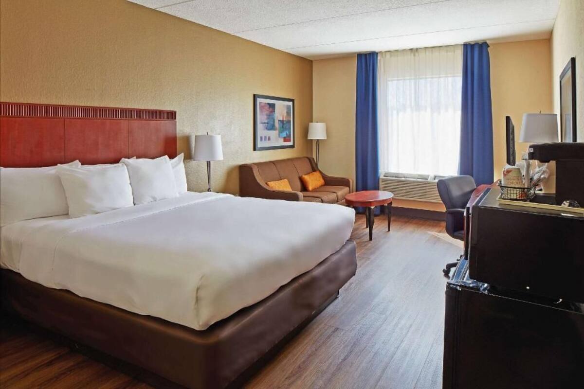A hotel room with a large bed, a desk, a chair, a small table, and a lamp.