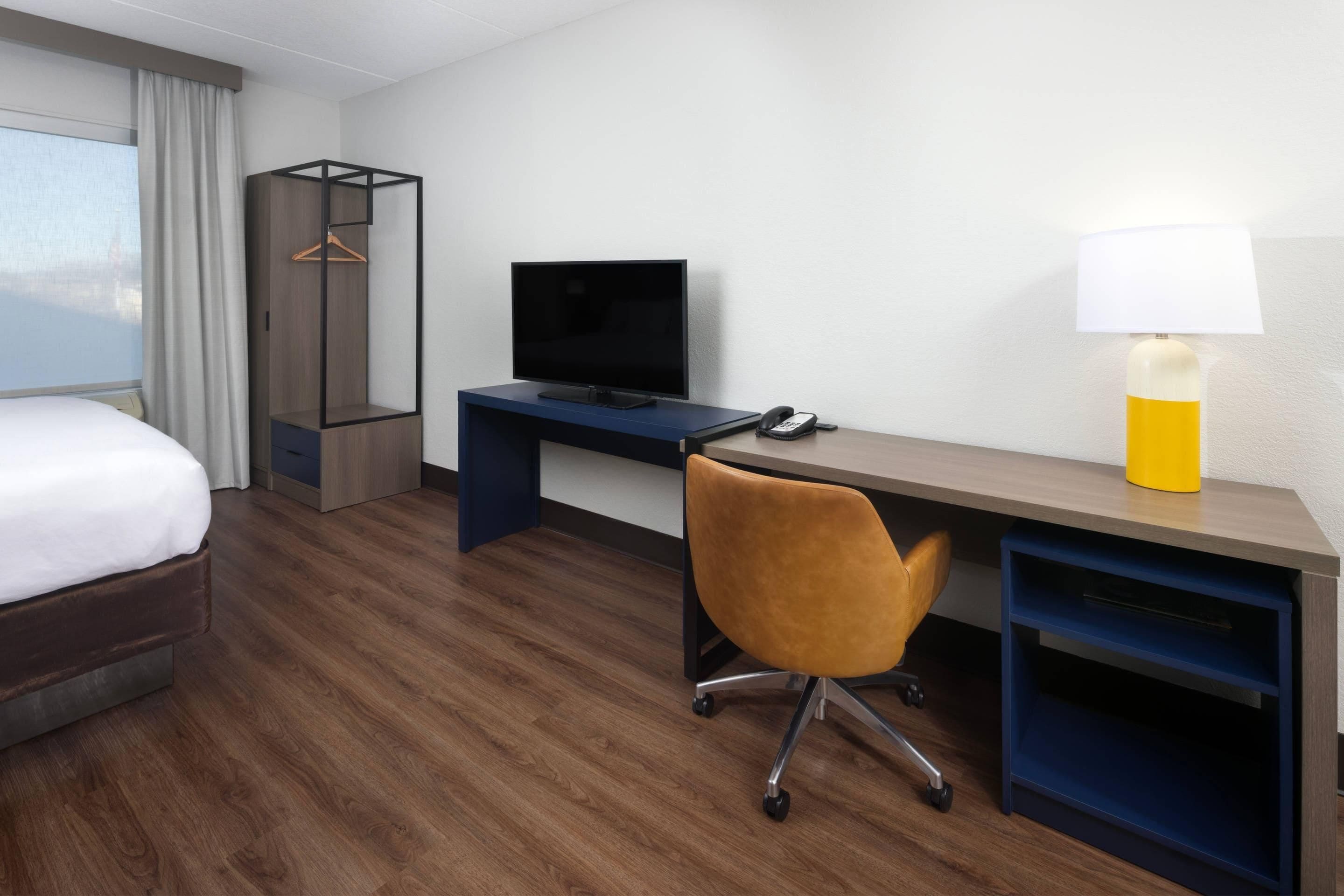 A hotel room with a bed, a desk with a TV, a chair, a lamp, and a wardrobe.