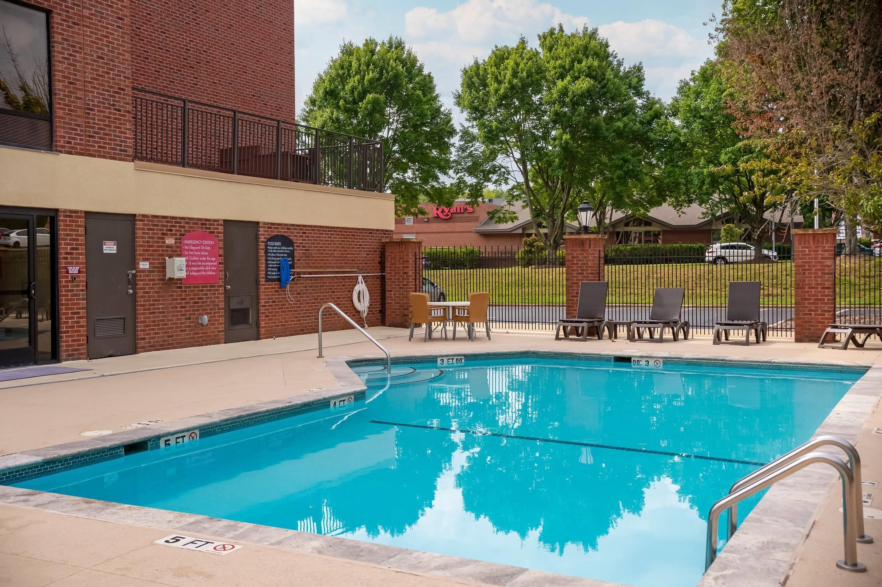 Una piscina con un edificio de ladrillo al fondo, una zona vallada con sillones de descanso y un letrero que anuncia el restaurante Ryan's.