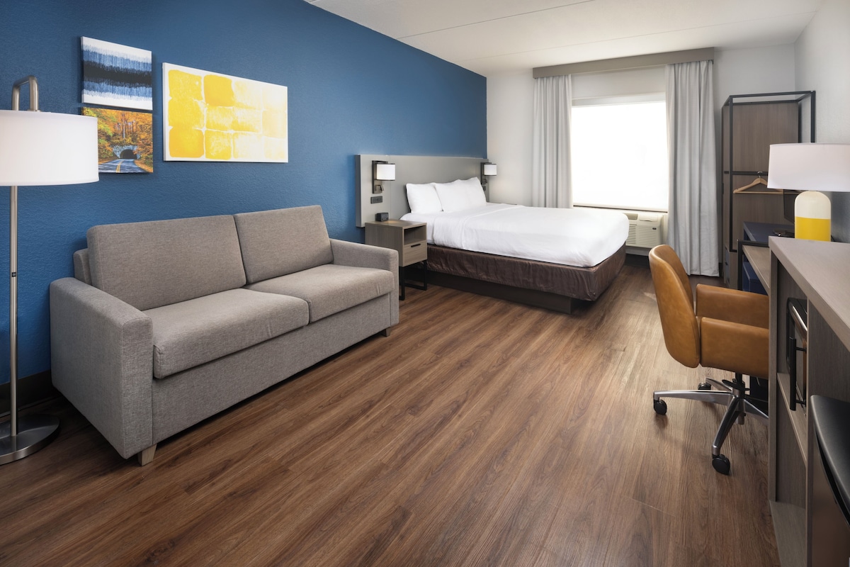 A hotel room with a bed, a sofa, a desk, and a chair. There are two bedside tables with lamps and a window with curtains.