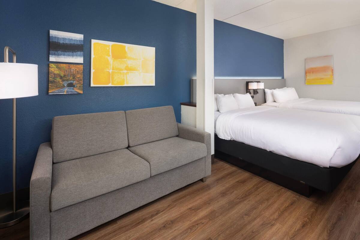 A hotel room with a grey sofa, a double bed with white bedding, a nightstand with a lamp, and artwork on the walls.