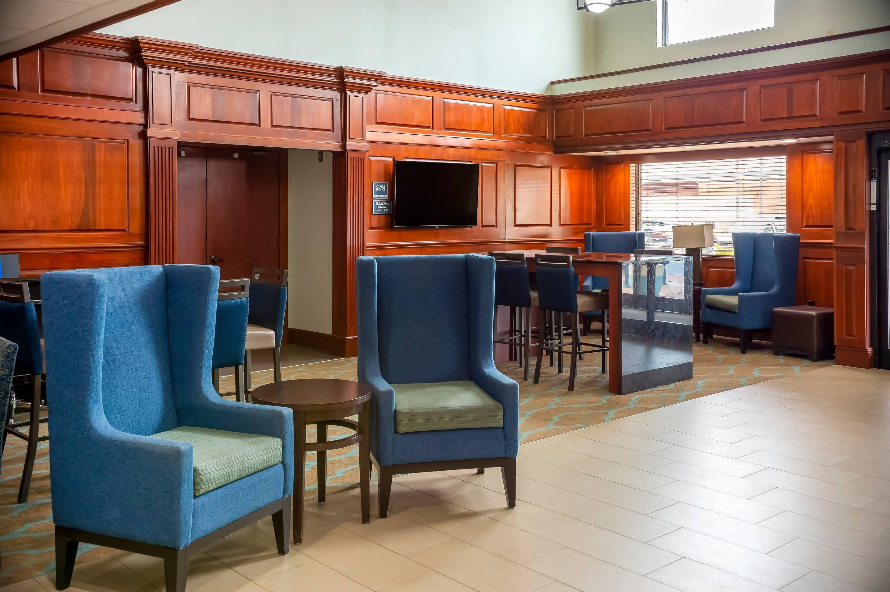 Un hall d’hôtel avec des boiseries, des fauteuils bleus, un panneau indiquant un service de café et un grand écran de télévision.
