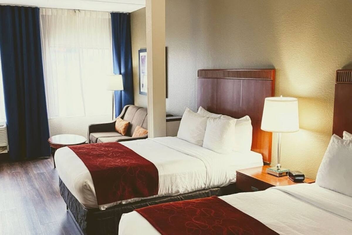 A hotel room with two beds, a small table, a chair, a lamp, and a window with curtains.
