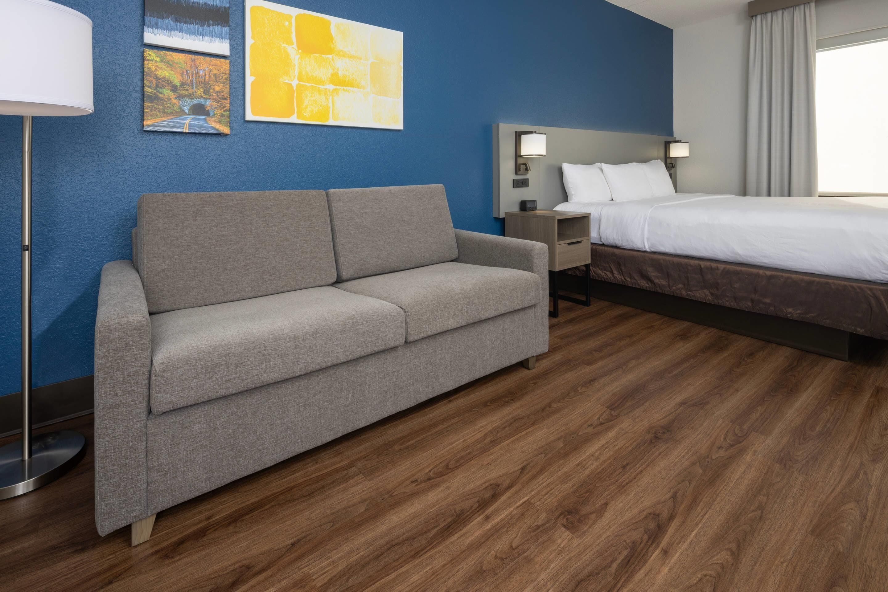 A hotel room with a grey sofa, a bed, a nightstand, and artwork on the walls.