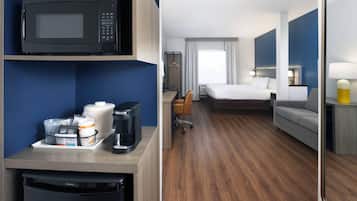 Habitación de hotel con microondas, cafetera y un refrigerador pequeño sobre una encimera. Se ve una cama, un sofá y un escritorio con una silla.