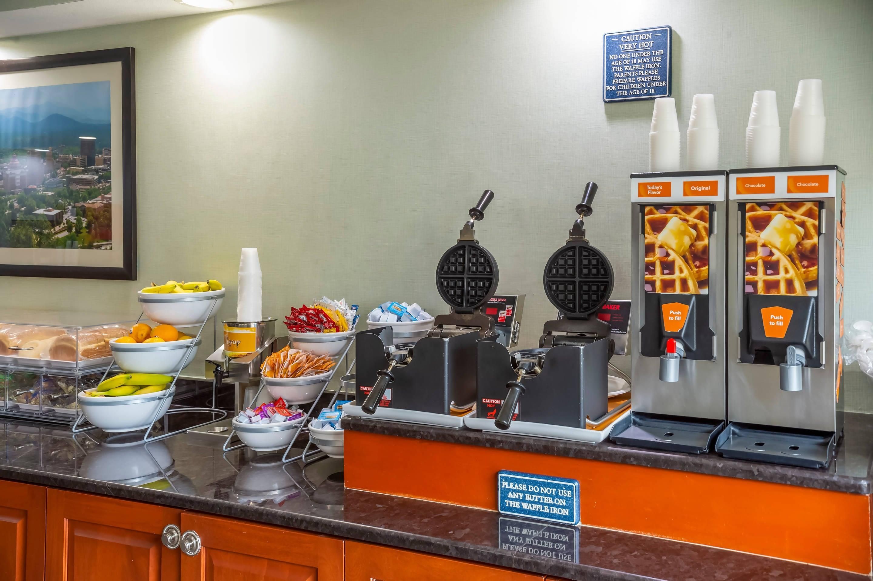 Un buffet de petit-déjeuner proposant des gaufriers, une variété de fruits, et un panneau d’avertissement interdisant d’utiliser du beurre sur le gaufrier.