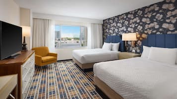 Habitación de hotel con dos camas, televisión, escritorio, silla y un amplio ventanal con vista a la ciudad.