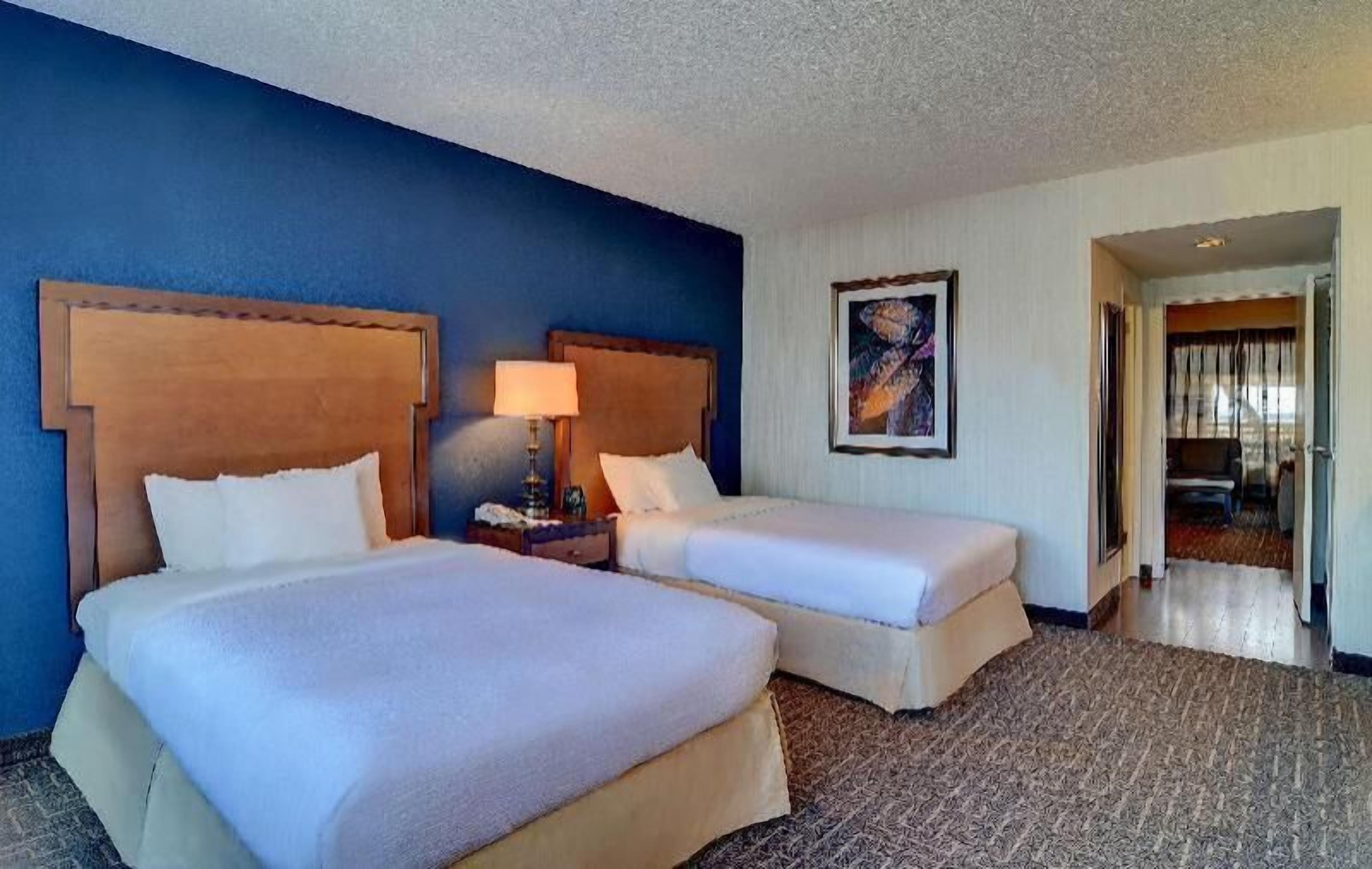 A hotel room with two beds, a nightstand with a lamp, a painting on the wall, and a view of the hallway.