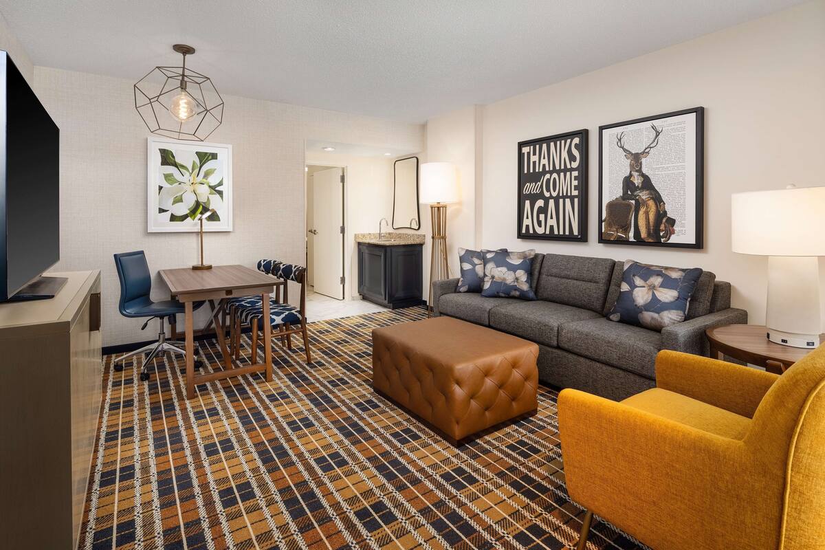 Et moderne hotelværelse med sofa, lænestol, skærm og spiseplads med bord og stole. Væggen er dekoreret med billeder i rammer, og der er en lampe.