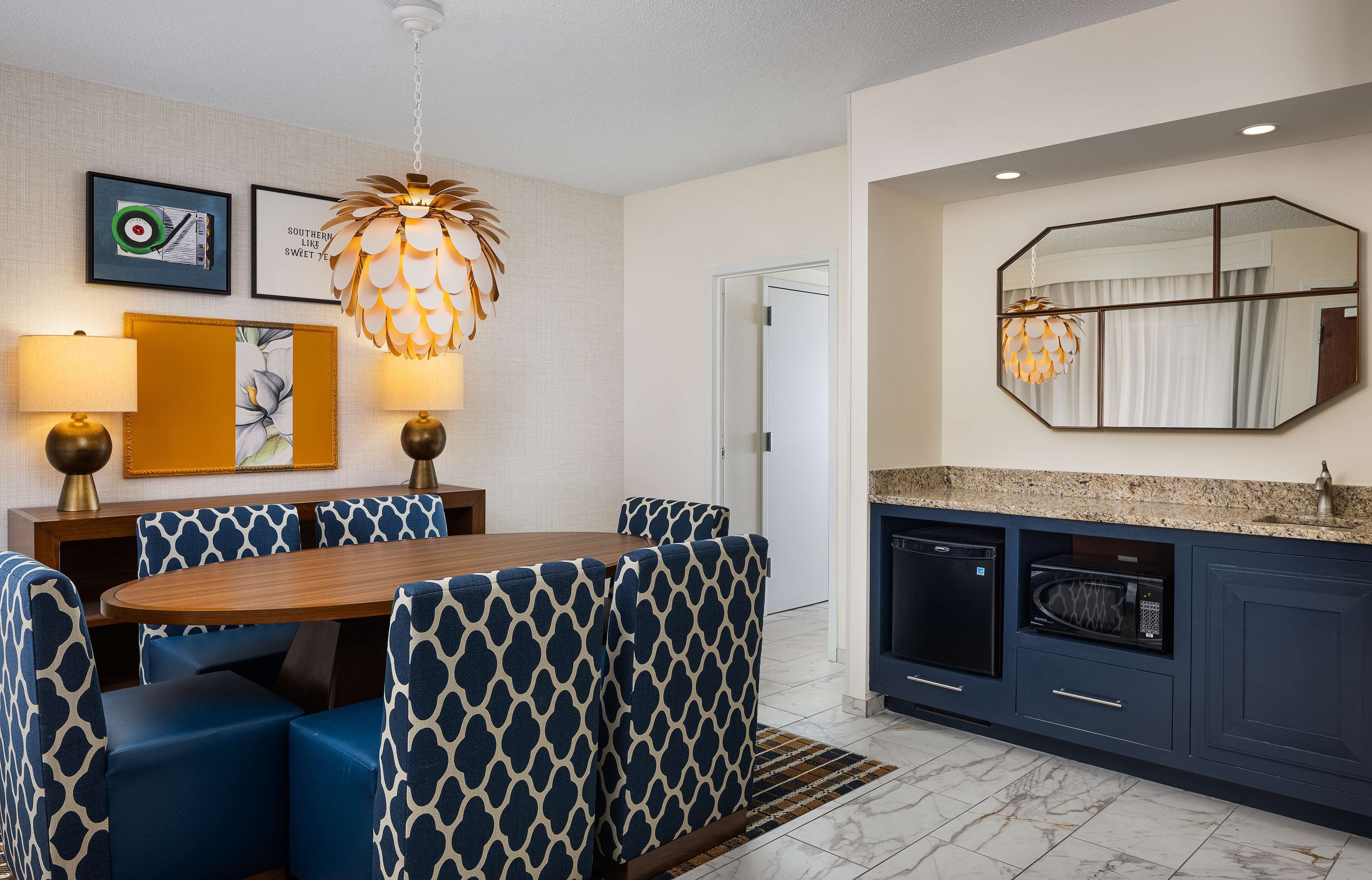 A modern kitchen with a dining area, featuring a wooden table, blue patterned chairs, a hanging light fixture, a microwave, and a large mirror.