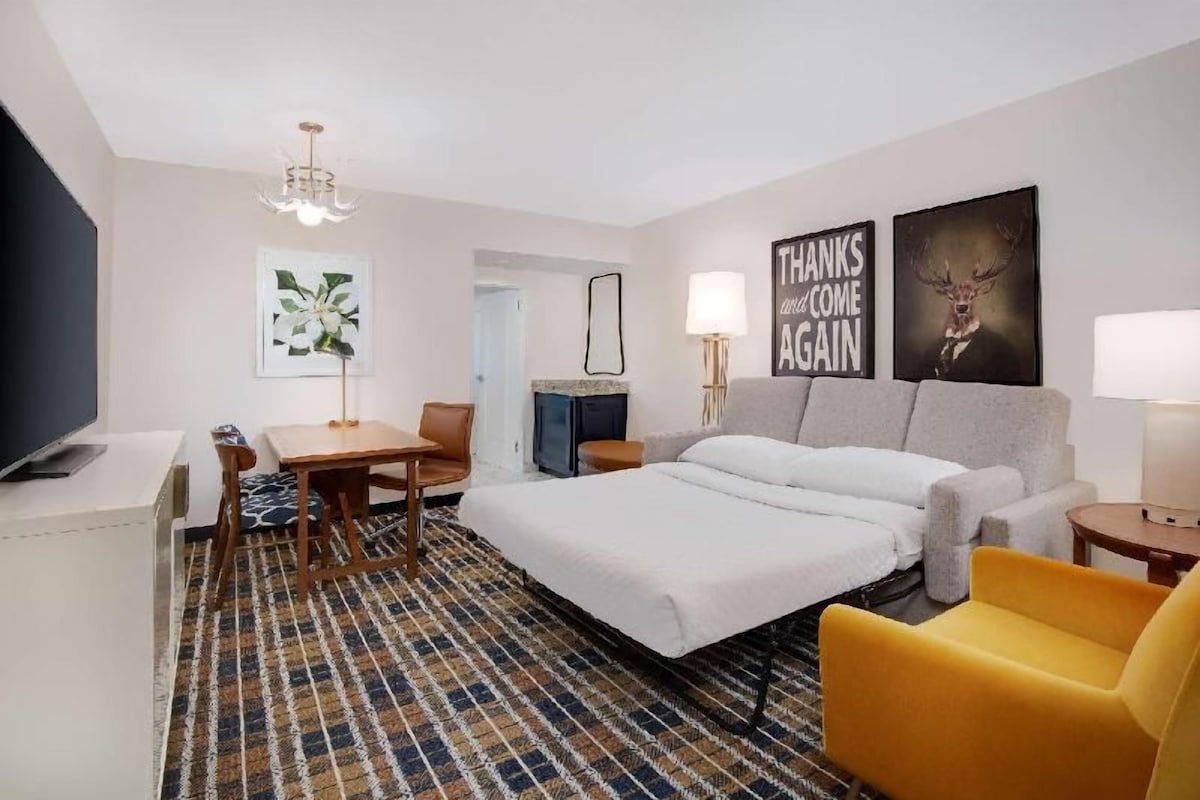 A hotel room with a bed, a desk, a chair, a sofa, a television, and artwork on the walls.
