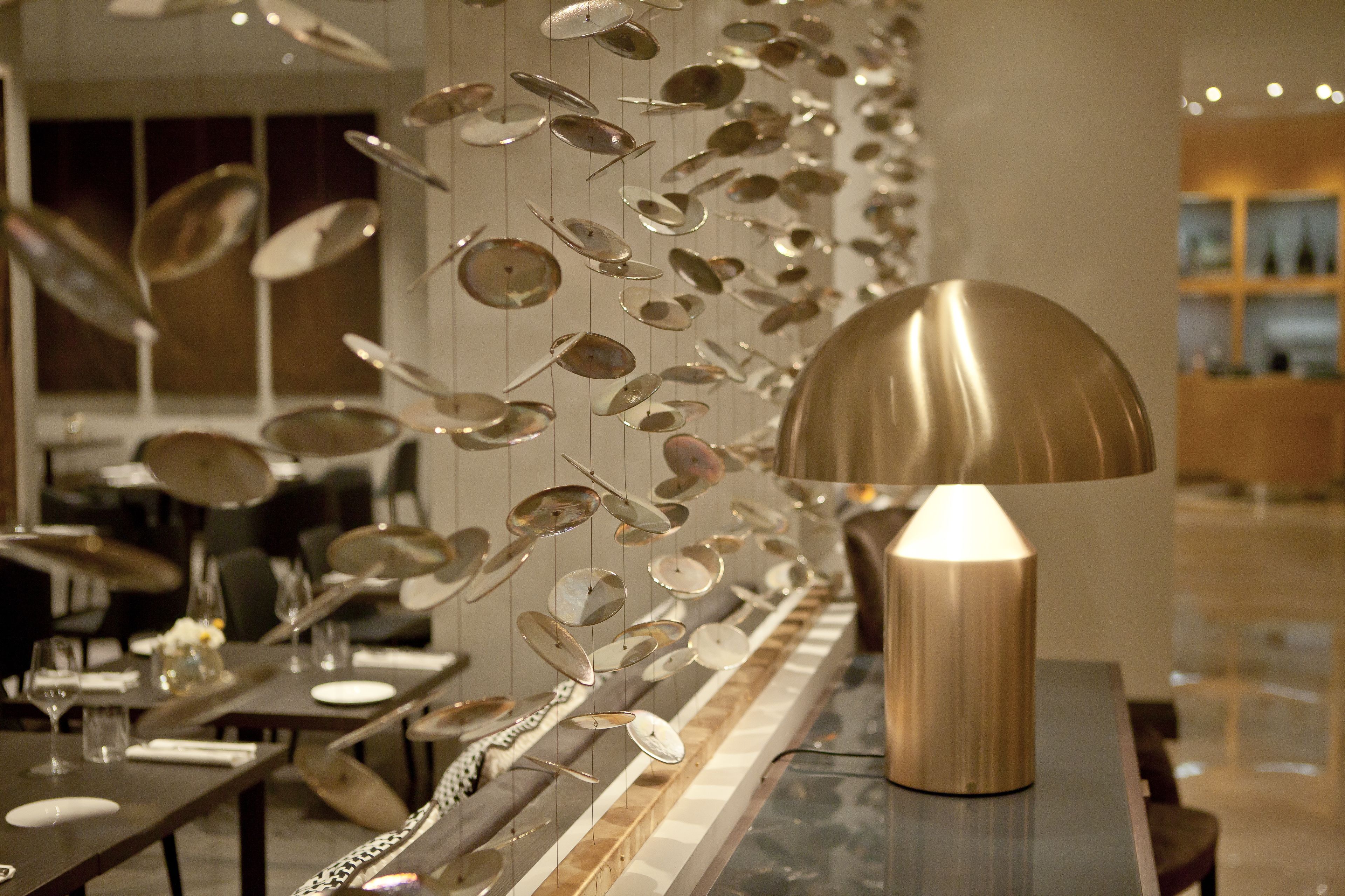 Ein moderner Essbereich mit einem langen Tisch, der für eine Mahlzeit gedeckt ist, einer Wand mit hängenden Metallobjekten und einer Strahlerlampe auf dem Tisch.