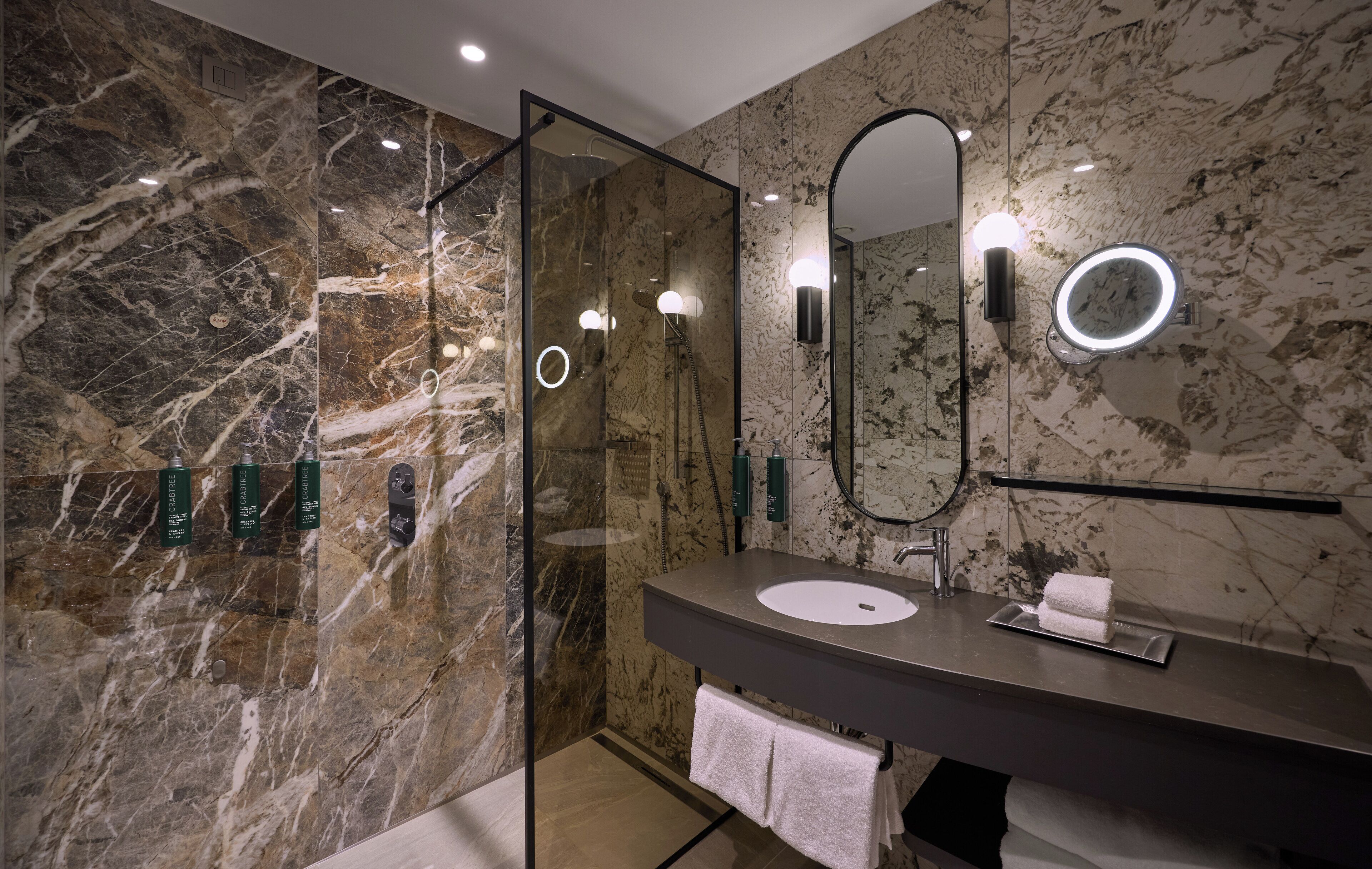 A modern bathroom with a marble wall, a glass shower enclosure, a round mirror with a light, and a dark countertop sink.