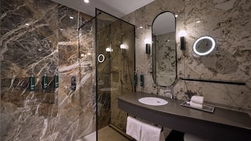 A modern bathroom with a marble wall, a glass shower enclosure, a round mirror with a light, and a dark countertop sink.