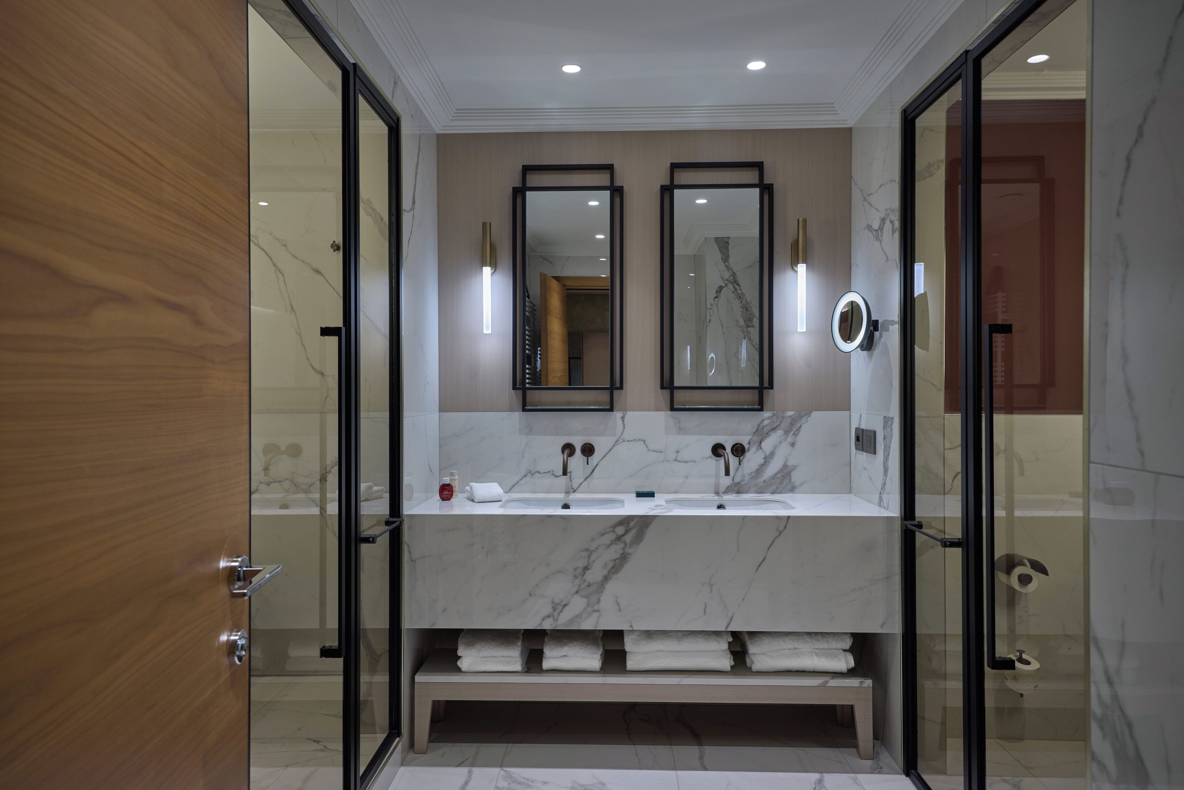 A modern bathroom with a marble vanity, two mirrors, a glass shower enclosure, and a wooden door.