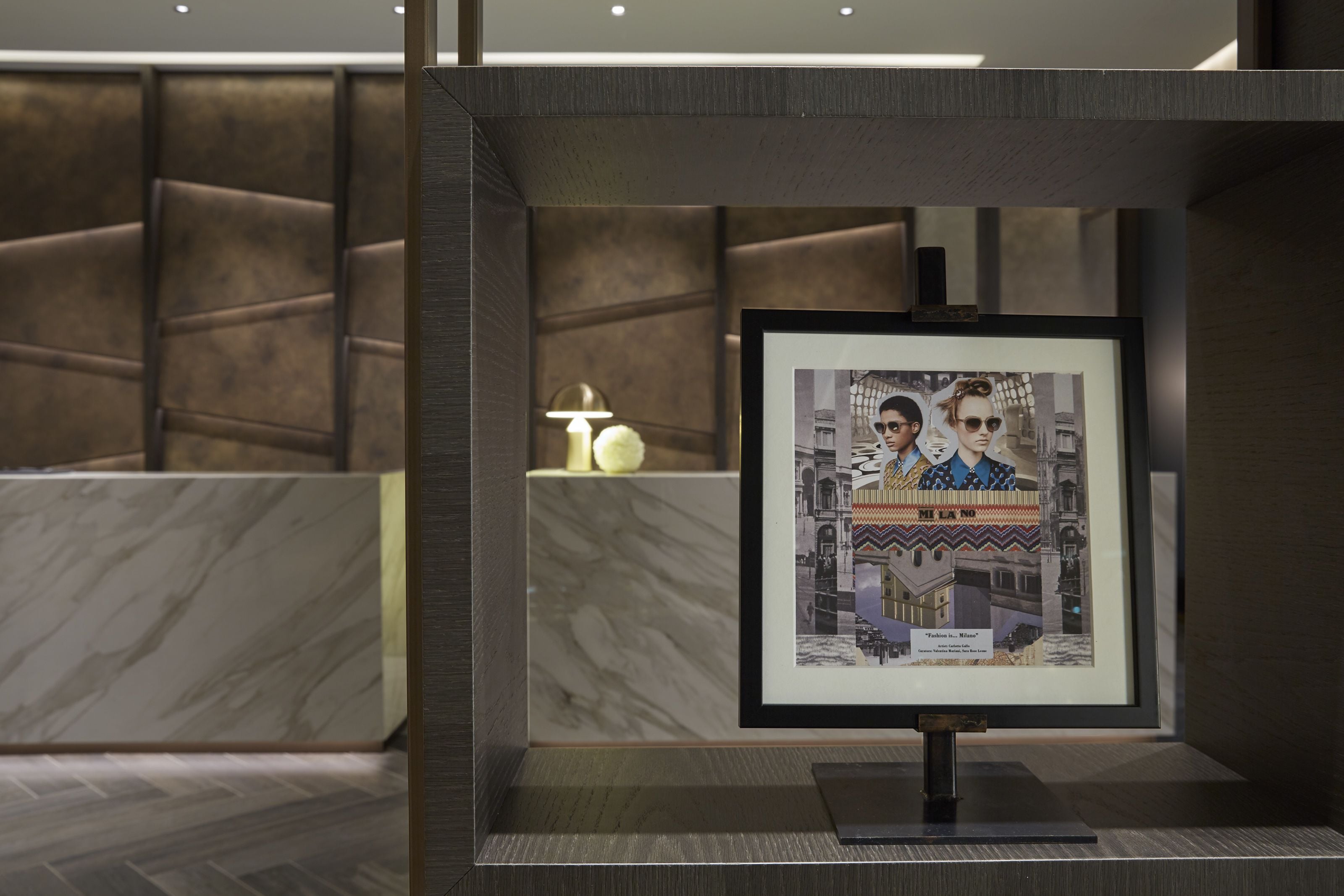 A framed picture of a building with a caption, placed on a stand in a modern interior with marble flooring and a patterned wall.