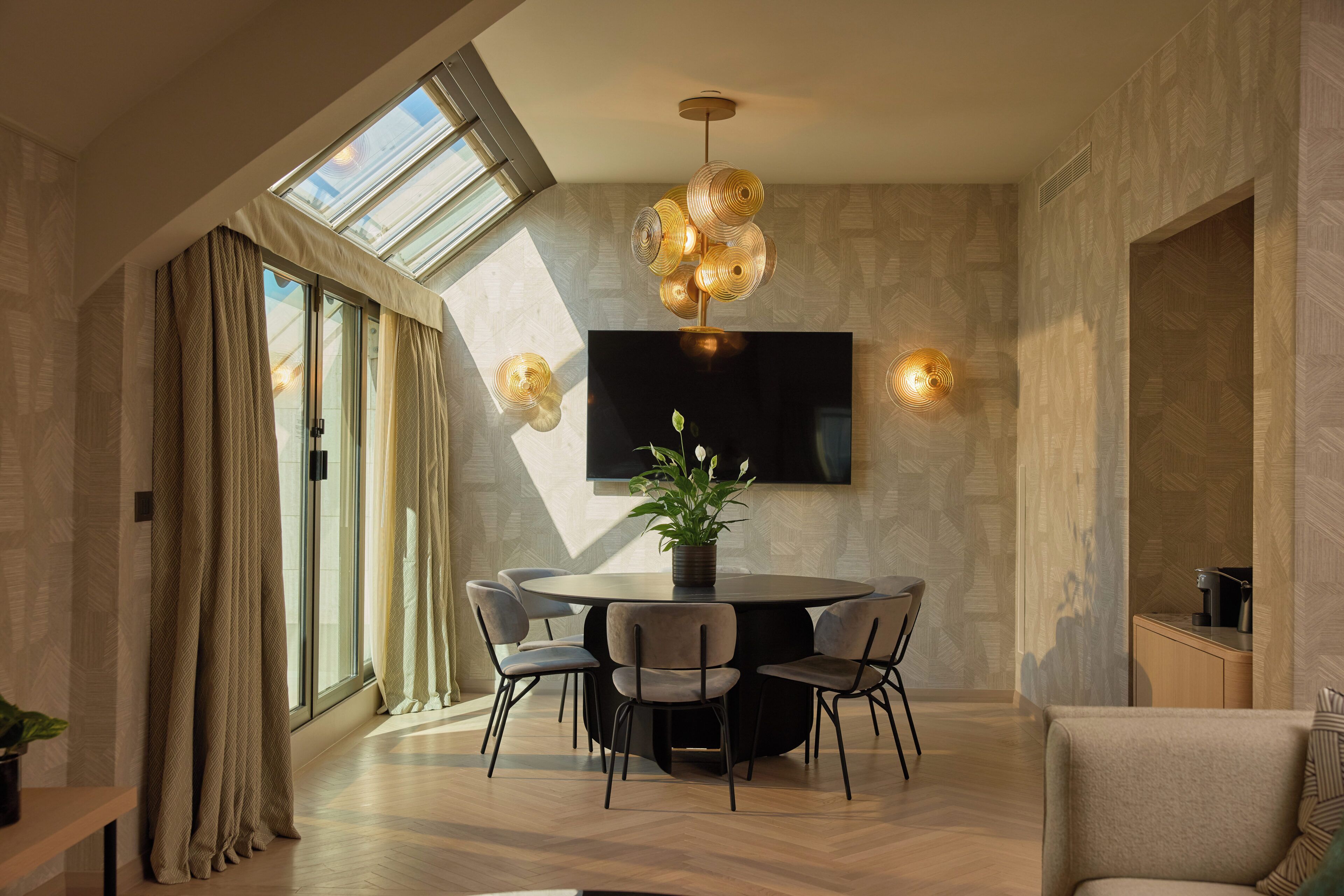 A modern dining area with a round table, chairs, a potted plant, a flat-screen TV, and a unique chandelier.