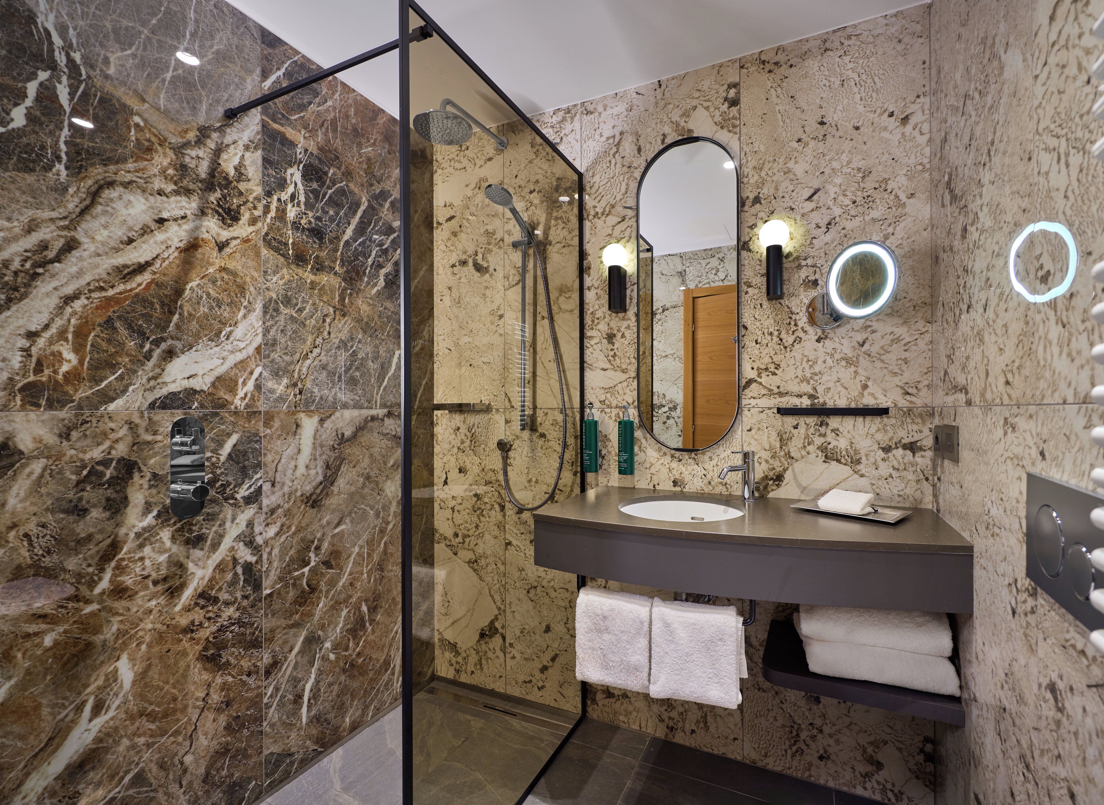 Une salle de bain moderne dotée d’une douche à parois de verre, de murs en marbre, d’un grand miroir et d’un meuble-lavabo avec un évier et un porte-serviettes.