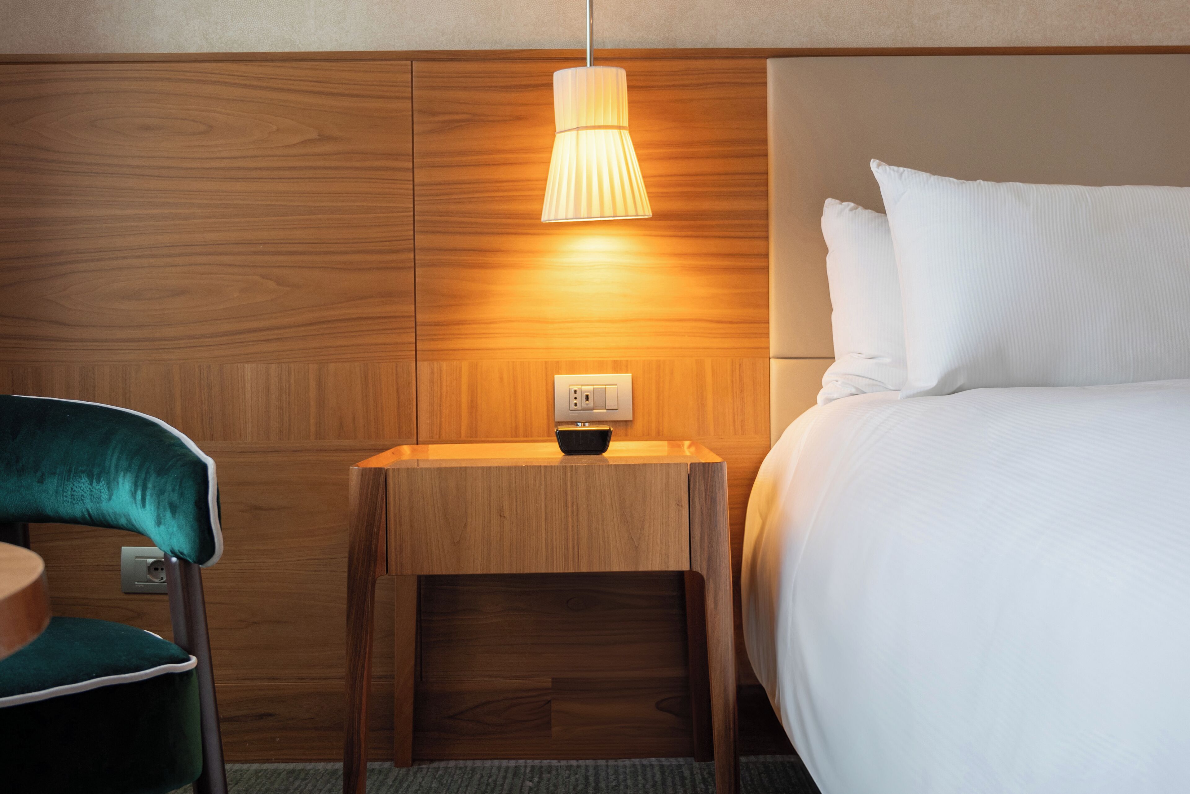 A hotel room with a wooden headboard, a bedside table with a lamp, a green chair, and a bed with white bedding.