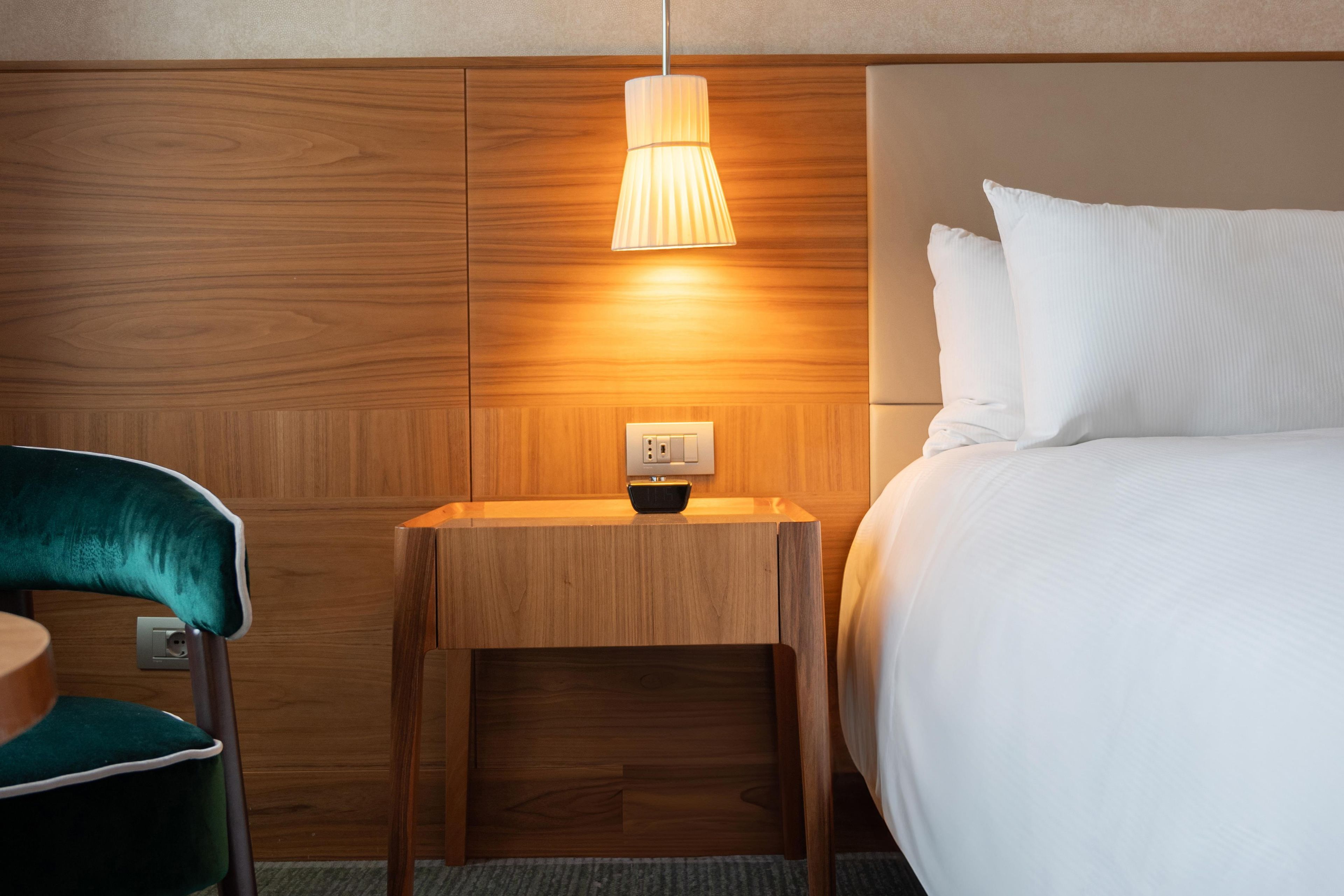 A hotel room with a wooden headboard, a bedside table with a lamp, a green chair, and a bed with white bedding.