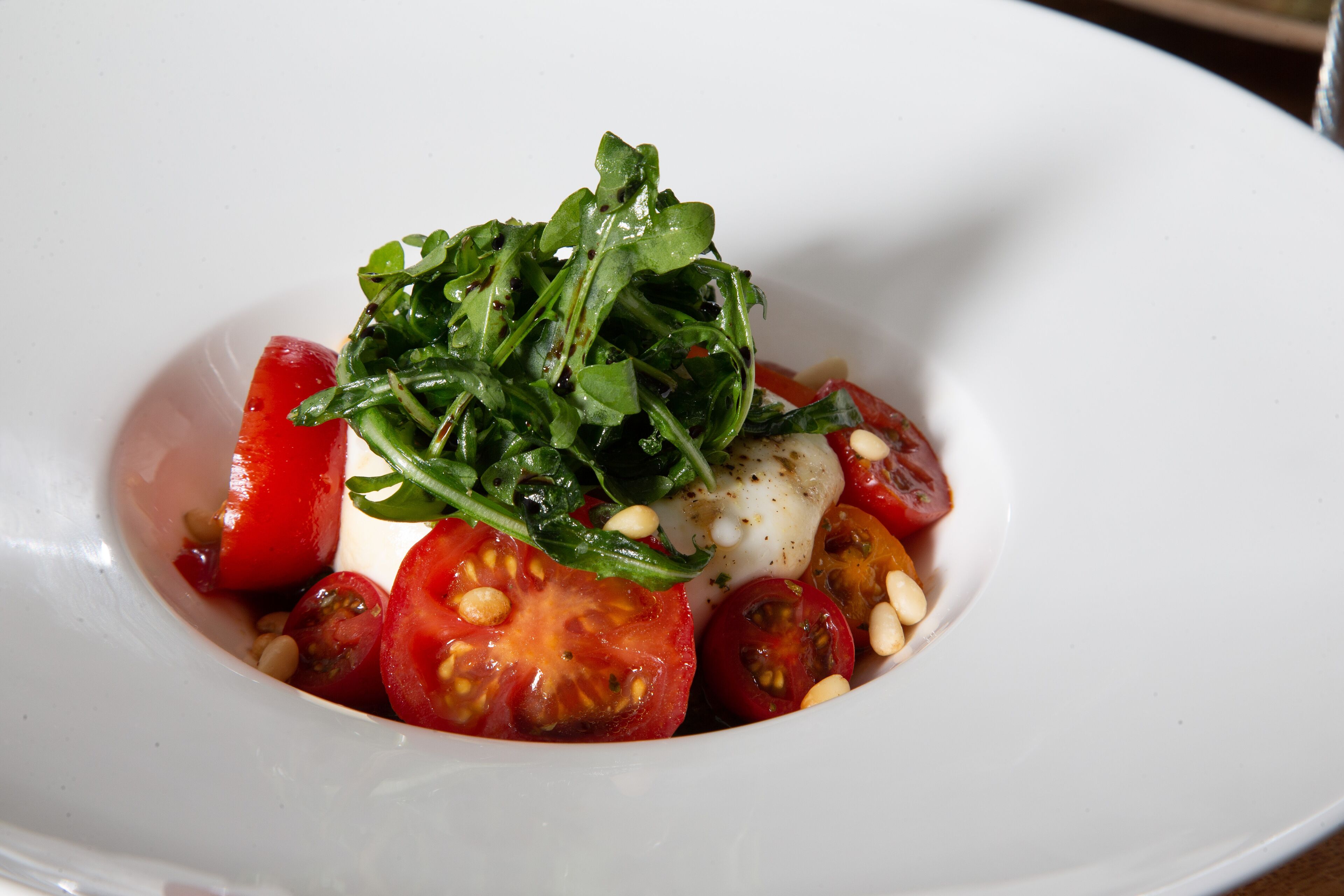 A white plate with a salad consisting of cherry tomatoes, mozzarella, arugula, and pine nuts.