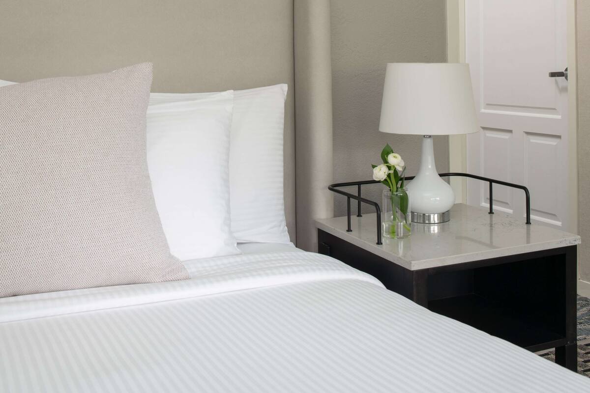 A neatly made bed with white linens and pillows, a bedside table with a white lamp and a vase with flowers, and a white door in the background.
