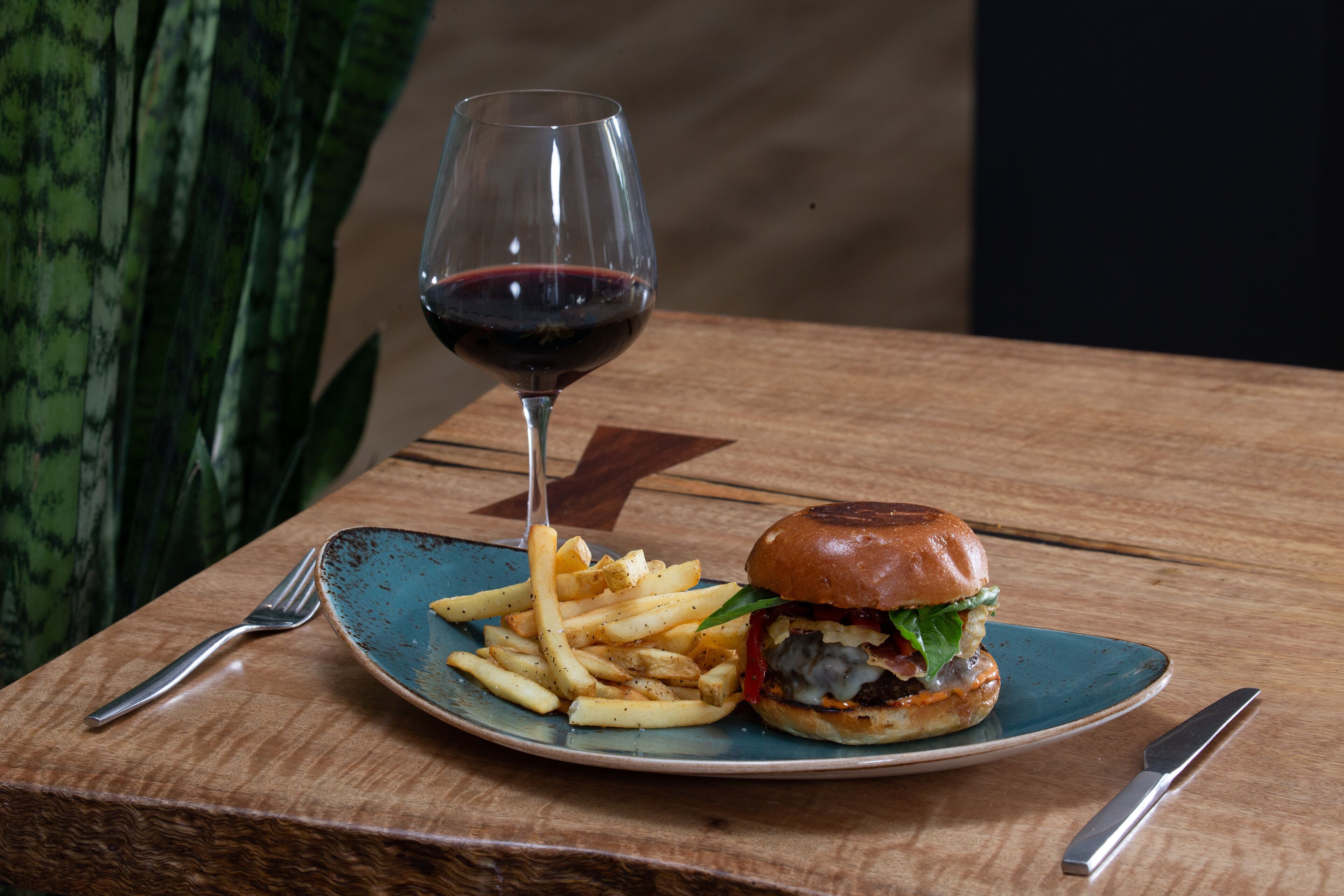 Een bord met een hamburger en frietjes, een wijnglas en bestek op een houten tafel.