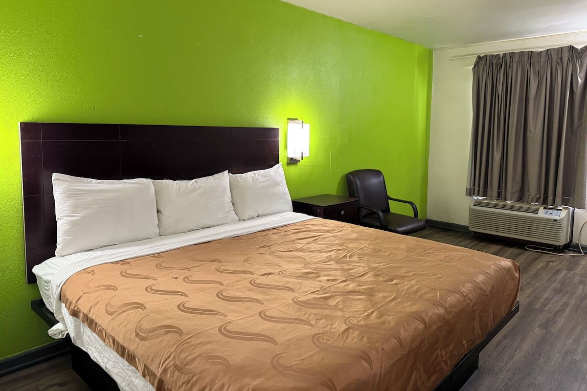 A hotel room with a bed, a desk, a chair, and an air conditioning unit.