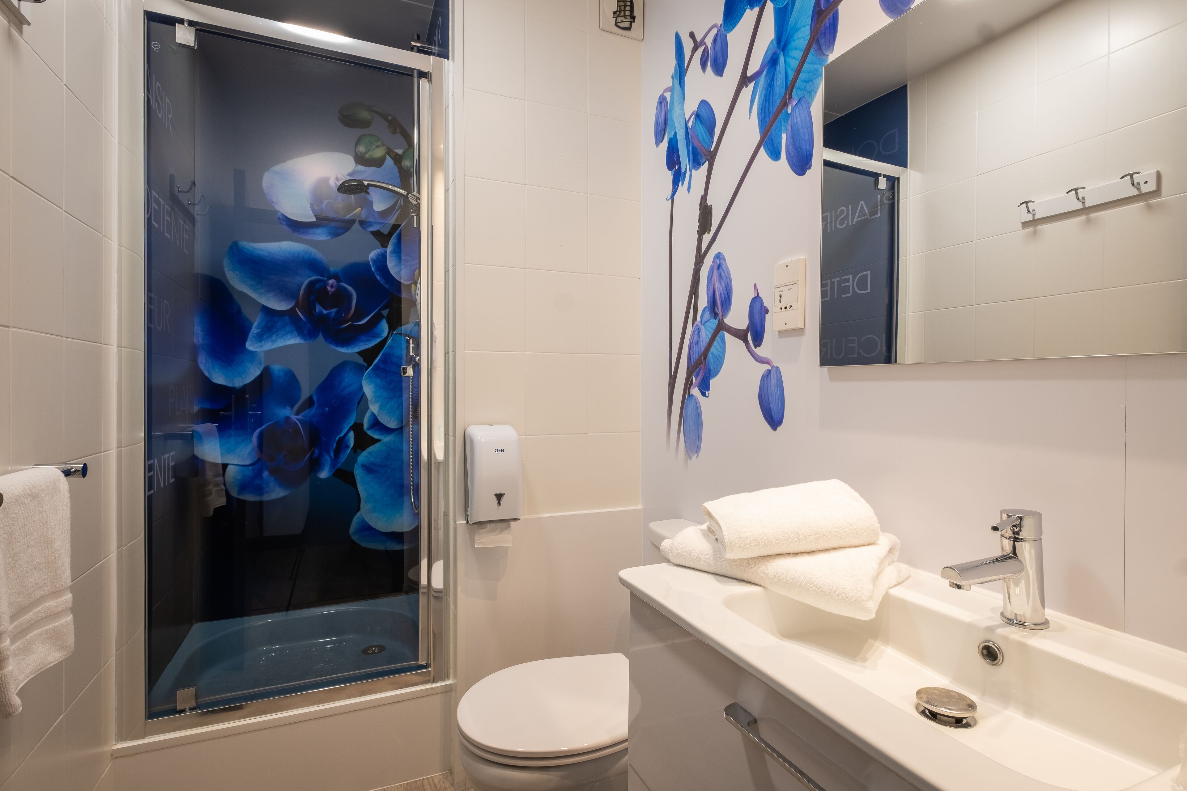 Une salle de bain avec une douche à parois de verre, des toilettes blanches, un lavabo avec un miroir et un mur orné d’une fresque représentant des orchidées bleues.