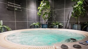 A hydro-massage bath with a control panel, surrounded by potted plants and a tiled wall with a sign.