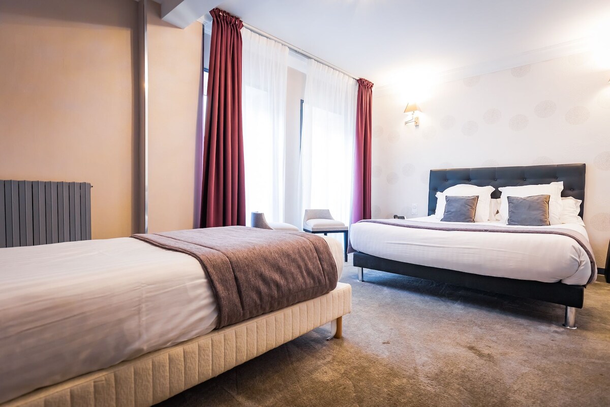 A hotel room with two beds, a chair, a radiator, and a window with curtains.