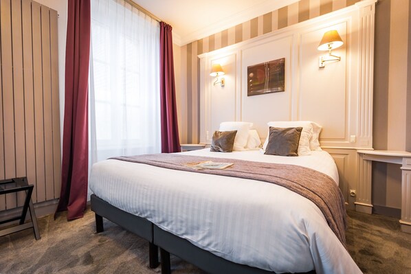 Quarto de hotel com uma cama grande, duas luminárias de cabeceira, uma janela com cortinas e um quadro na parede.