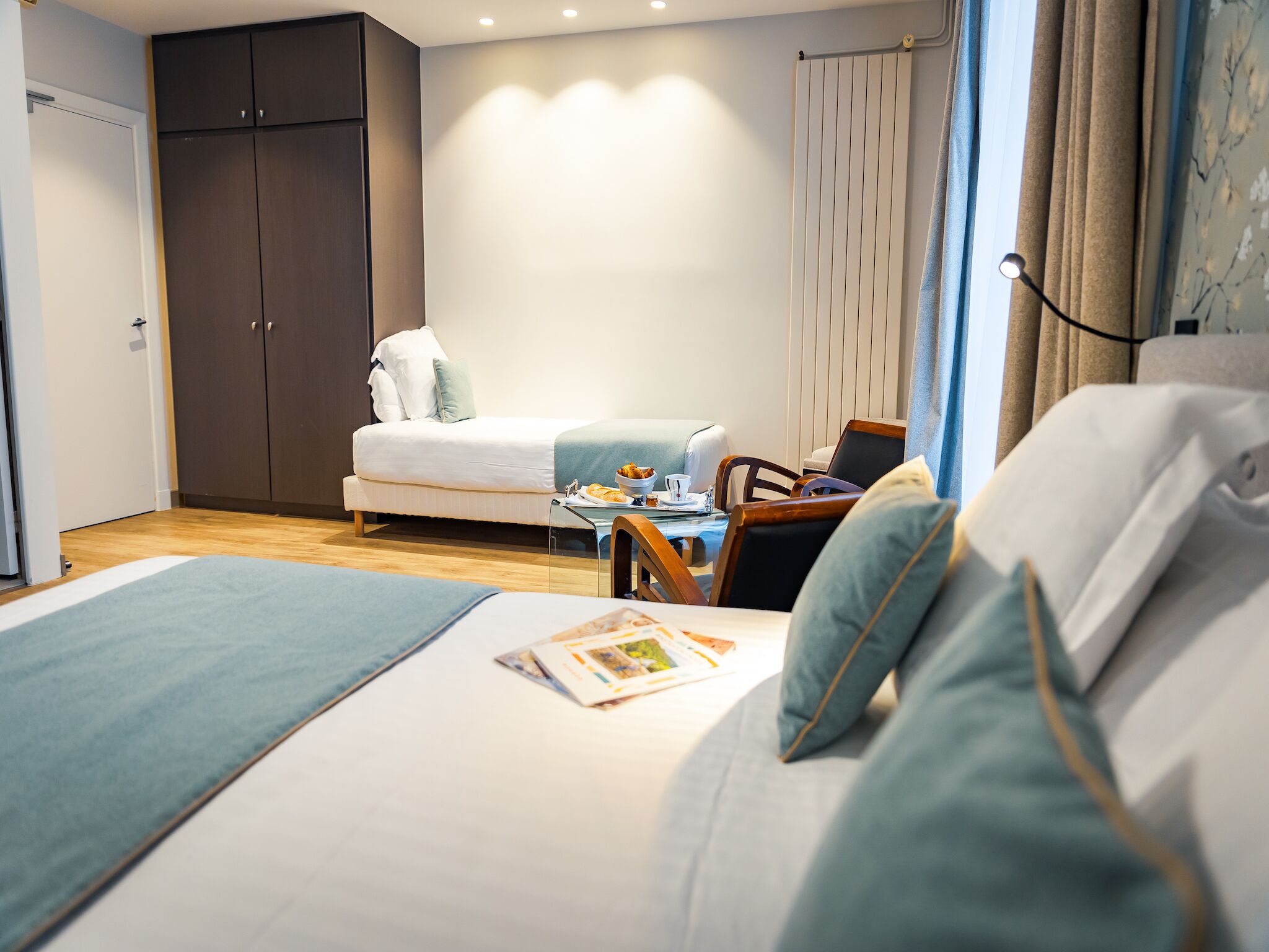 A modern hotel room with a bed, a small table with breakfast, a wardrobe, and a window with curtains.