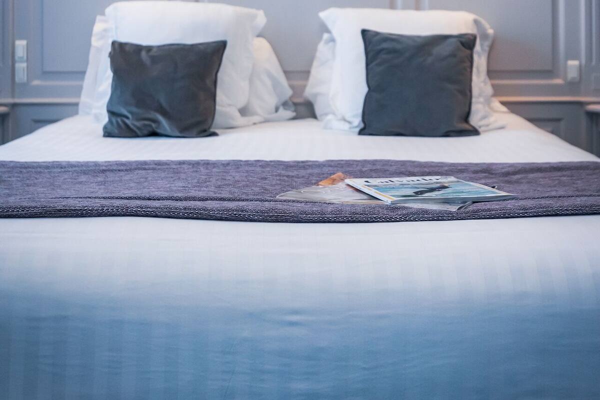 Un lit bien fait, avec une couverture violette, deux oreillers et un magazine posé sur le lit.